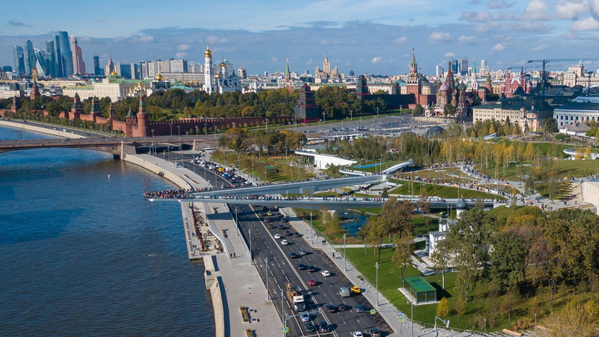 Obrázek je letecký pohled na Moskvu s řekou Moskvou, kremelskými zdmi z červených cihel, katedrálou Vasila Blaženého a moderními mrakodrapy, jako jsou ty v Moskvě. Ukazuje rušný park podél břehu řeky, mosty a podzimní listí pod částečně zataženou oblohou, kde se mísí historické a současné prvky města.