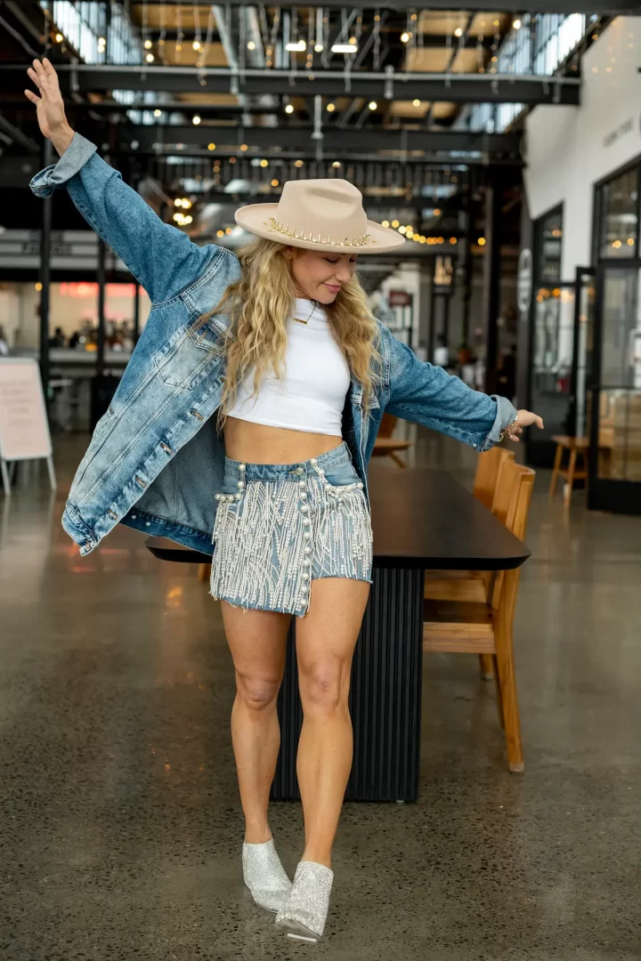 Žena s dlouhými vlnitými blond vlasy tančí v industriálním prostoru s vysokým stropem a světýlky. Má na sobě bílý crop top, džínovou bundu, krátkou džínovou sukni s perlovými třásněmi, třpytivé boty na podpatku a béžový klobouk se zlatými detaily.