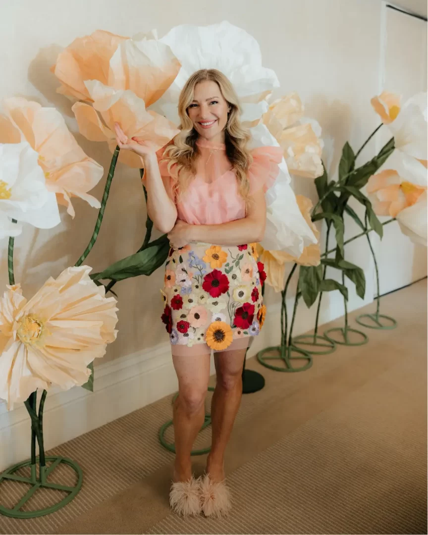 Usměvavá žena s dlouhými blond vlasy pózuje před aranžovanými obřími papírovými květy v odstínech bílé, krémové a broskvové. Má na sobě růžovou průsvitnou halenku s volánovými rukávy, krátkou sukni s 3D květinovou výšivkou a nadýchané chlupaté pantofle.