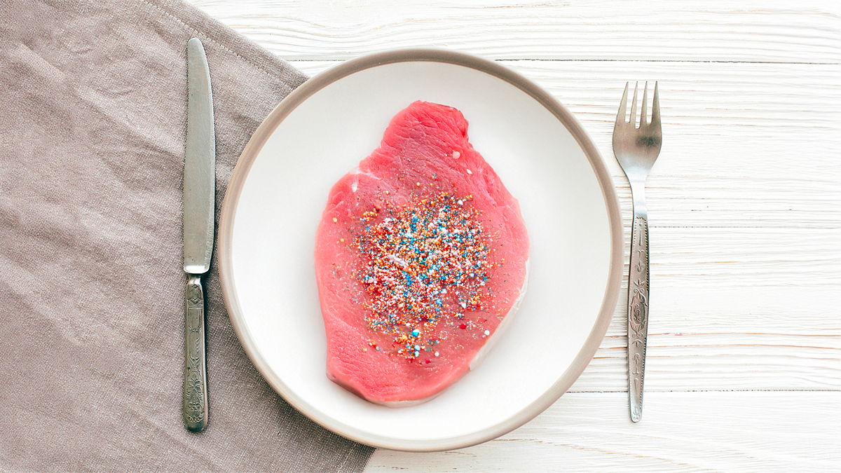 Syrový steak na talíři posypaný barevnými mikroplastovými částicemi, symbolizující přítomnost mikroplastů v potravinách. Vedle talíře je nůž a vidlička na dřevěném stole.