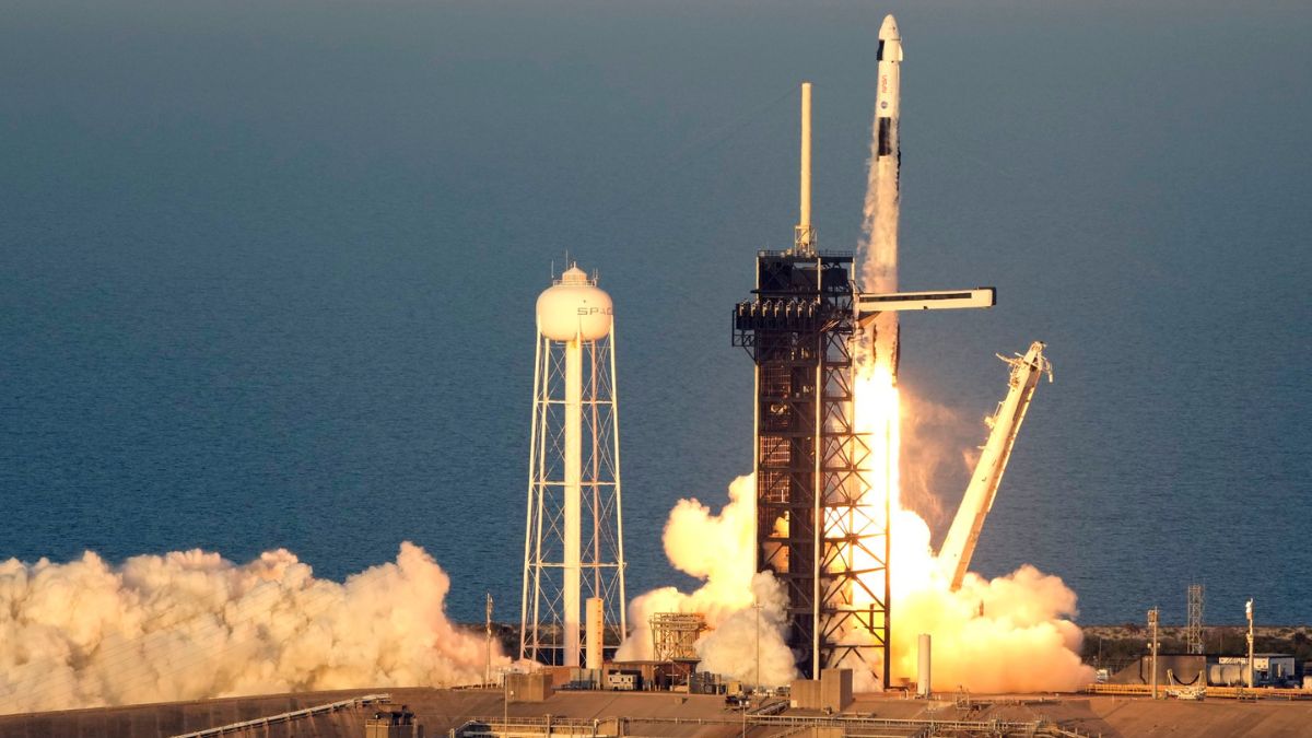 Obrázek znázorňuje probíhající start rakety. Raketa, která se podle svého designu a označení na vodárenské věži jeví jako SpaceX Falcon 9, se zvedá z odpalovací rampy. Raketa je obklopena velkým oblakem kouře a plamenů, když se její motory zapalují, což vytváří dramatickou a dynamickou scénu. Odpalovací věž se svou strukturou je vidět napravo od rakety a raketa právě opouští věž, když stoupá.