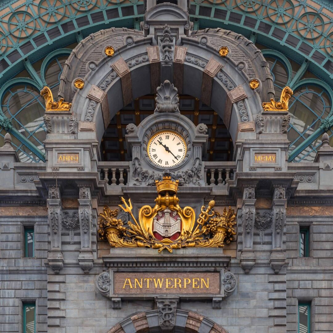 Detail hodin a městského znaku na průčelí nádraží Antwerpen-Centraal, obklopený kamennými ornamenty a pozlacenými dekoracemi.