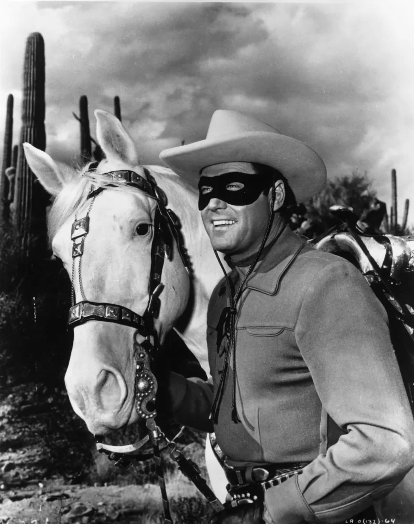 Maskovaný muž v klobouku stojí vedle bílého koně v pouštní krajině s kaktusy.